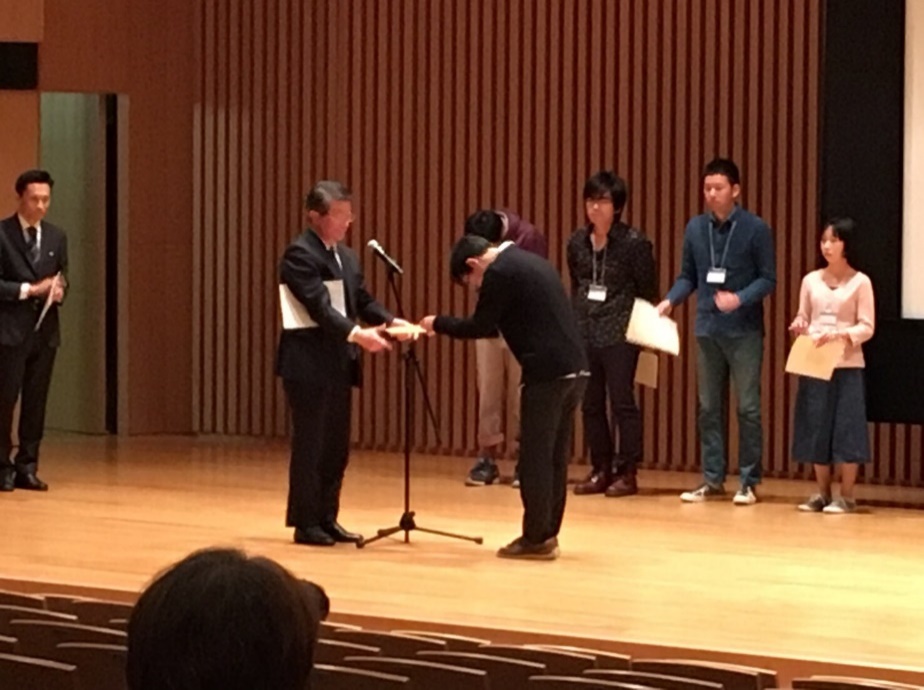 写真：深田吉孝先生 （東京大学・教授：日本時間生物学会次期理事長）から表彰状を受けとる水田さん（名古屋大学豊田講堂）。