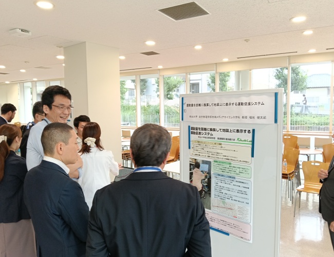 総合数理学部 先端メディアサイエンス学科の福地健太郎教授（写真左）による研究「運動量を距離に換算して地図上に表示する運動促進システム」のディスカッション時の様子